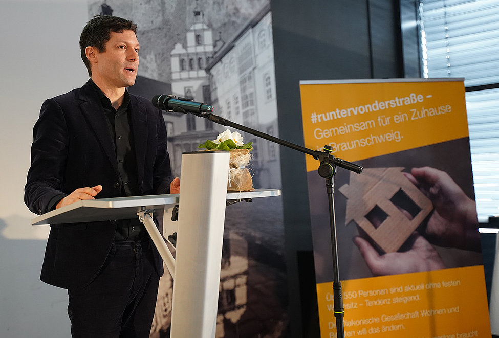 Ein Mann spricht am Rednerpult, daneben ein gelbes Plakat zur Aktion „#runtervonderstraße – Gemeinsam für ein Zuhause in Braunschweig“.