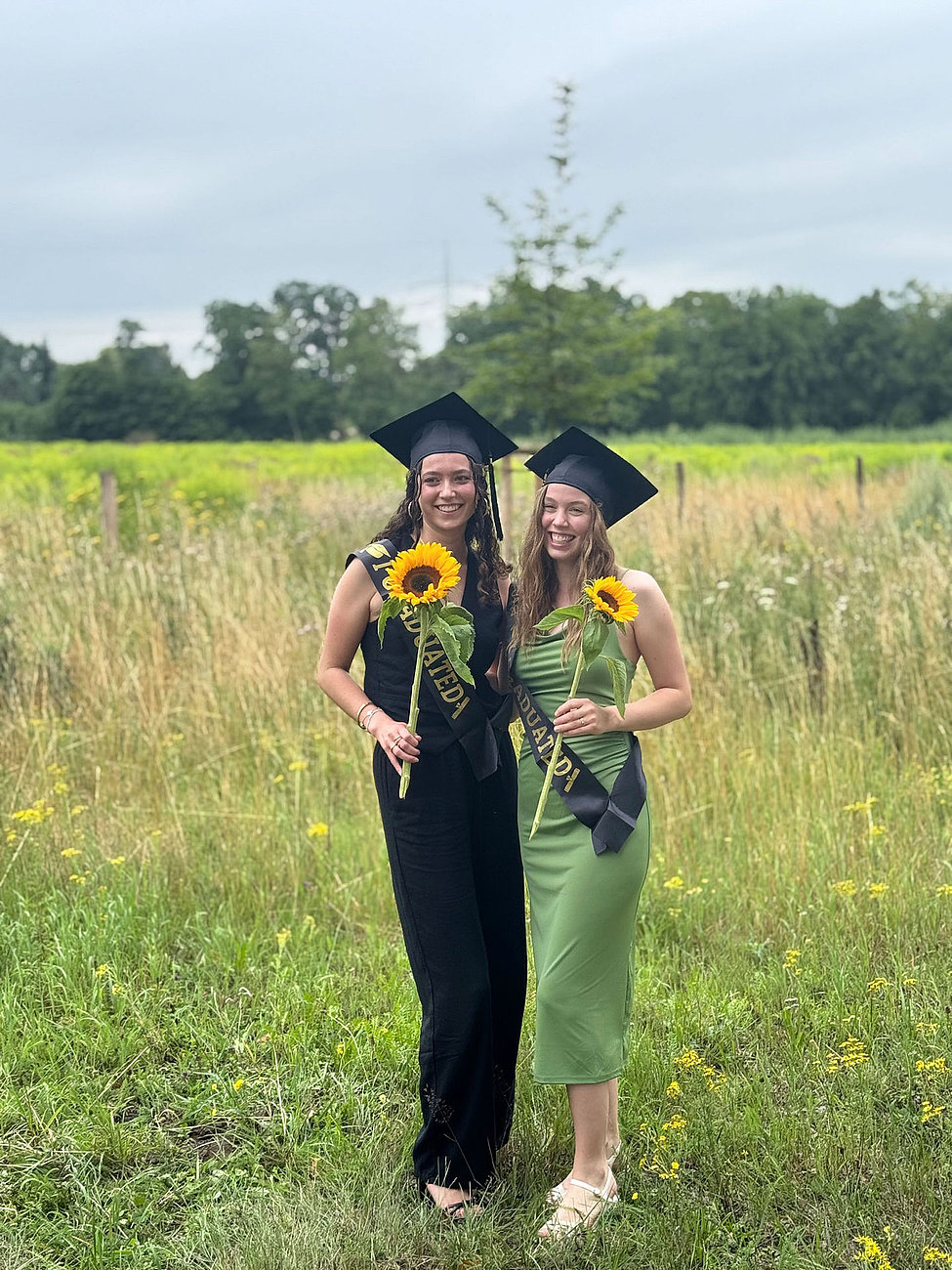Zwei Absolventinnen des dualen Studiums mit Sonnenblumen und Abschlusshüten auf einer Wiese