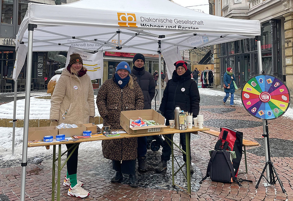 Vier Menschen stehen an einem Infostand unter einem Pavillon in der Innenstadt. Auf dem Tisch liegen kleine Giveaways, daneben steht ein Glücksrad.